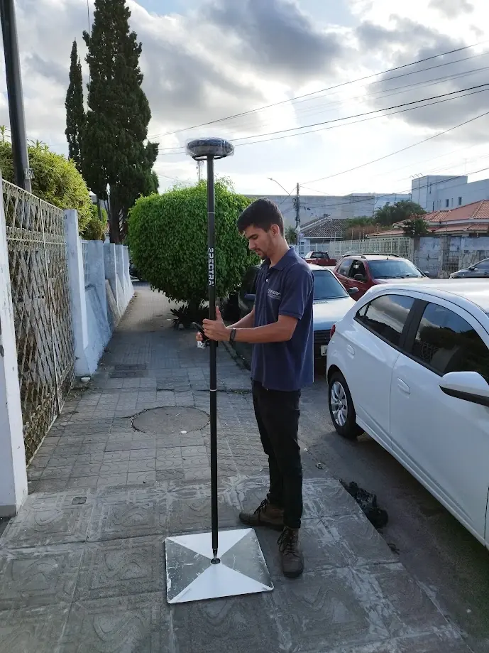 Ralph Araujo com bastão e receptor GNSS sobre uma base metálica numa calçada de rua urbana