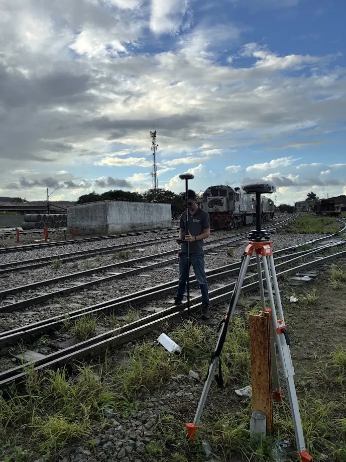 Técnico com receptor GNSS sobre os trilhos de um pátio ferroviário, com locomotivas ao fundo e receptor base em tripé no primeiro plano