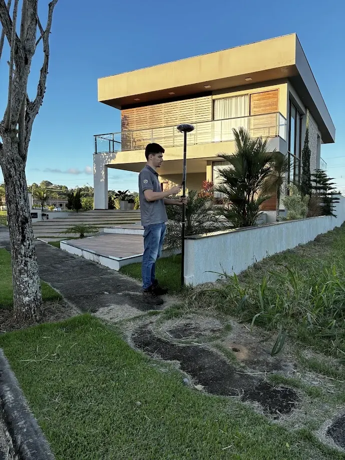 Ralph Araujo com receptor GNSS e coletora diante de uma casa de dois pavimentos, em lote urbano gramado
