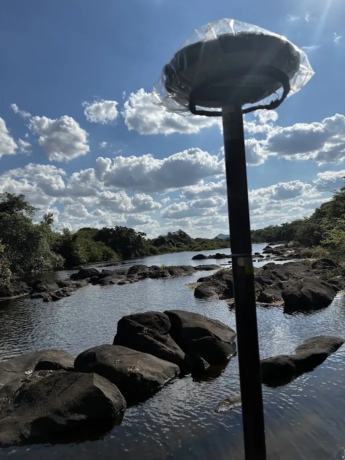 Antena GNSS em primeiro plano sobre o leito de um rio raso com rochas escuras e mata ciliar