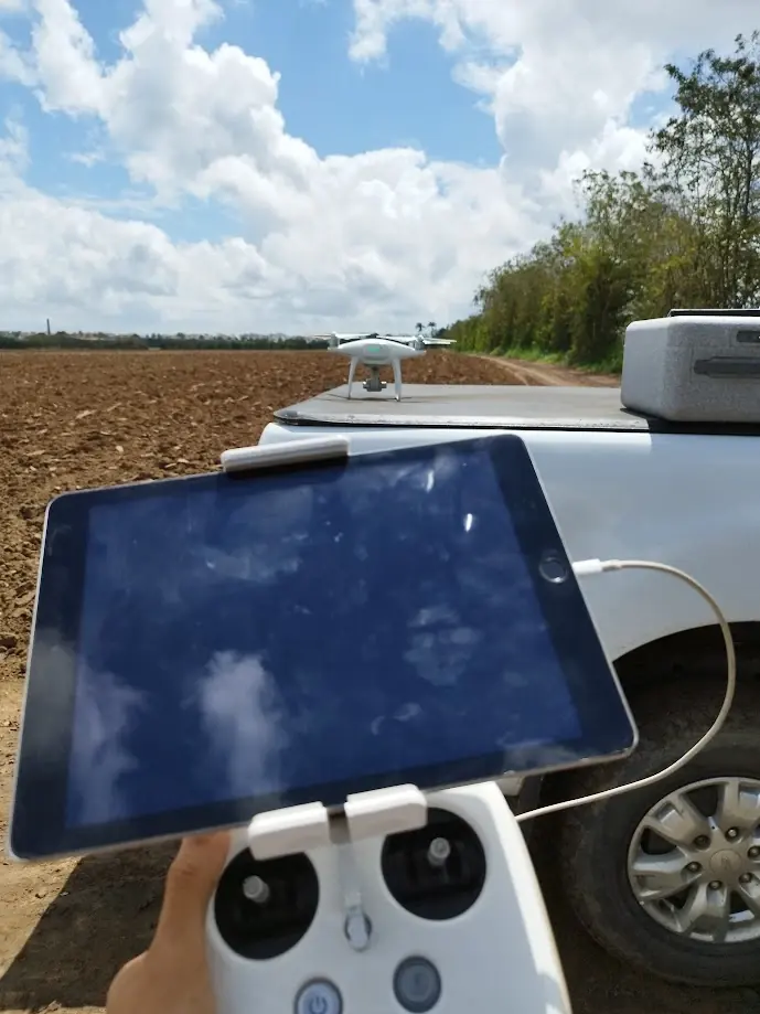 Drone pousado sobre a caçamba de uma caminhonete, à beira de uma área de solo recém-preparado, com o controle e o tablet em primeiro plano